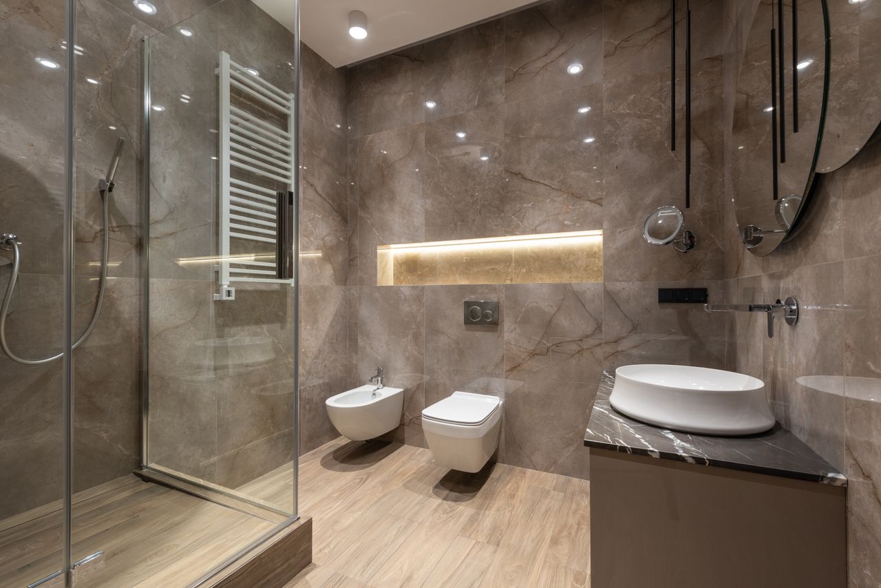 services-01 Glass shower cabin and ceramic sink with toilet bowl and bidet in spacious contemporary bathroom with shiny tiles and round mirrors